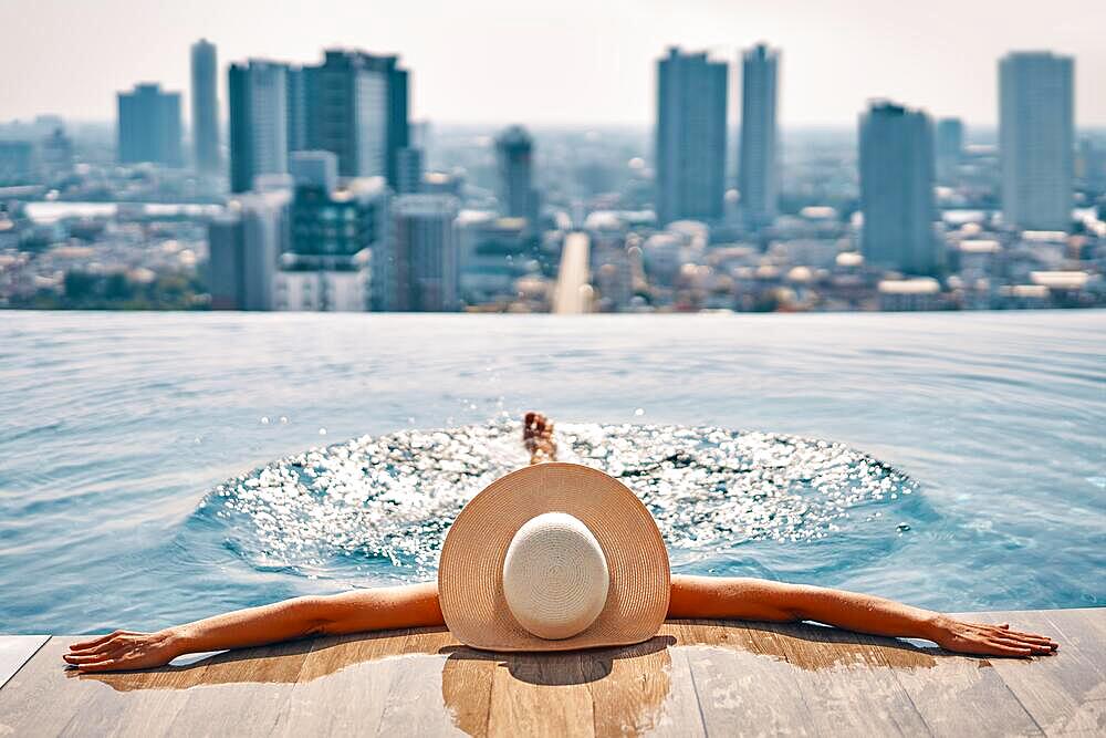 Back view of young woman in hat relaxing in swimming pool on the roof top of hotel and enjoy cityscape. Summer vacation, rest concept