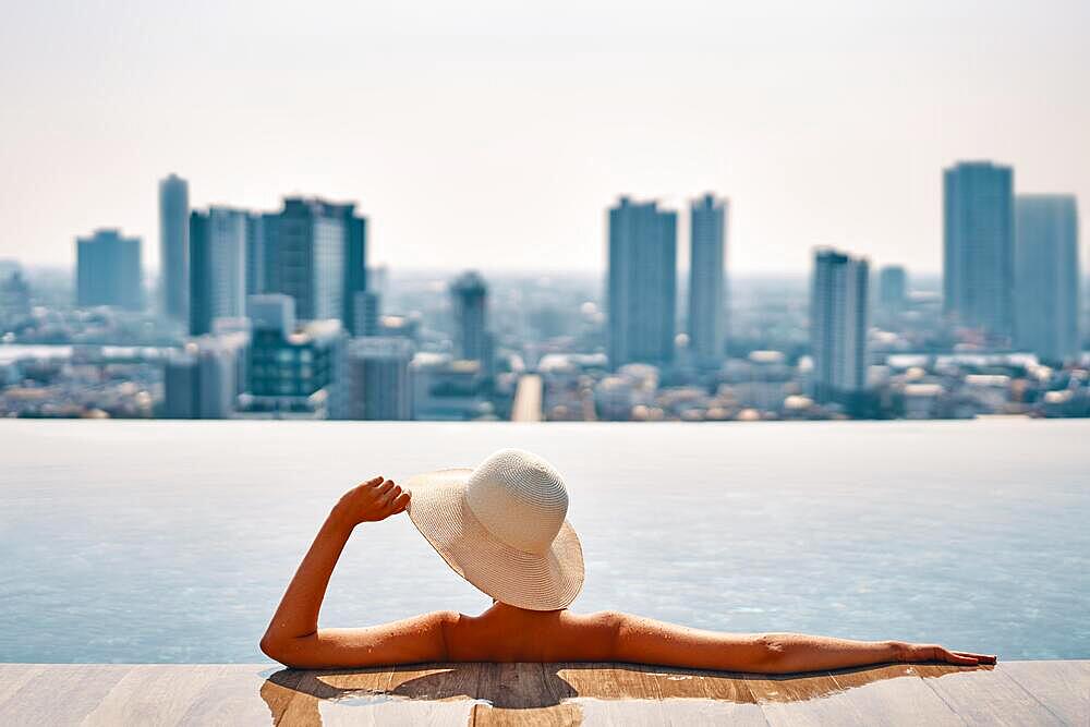 Back view of young woman in hat relaxing in swimming pool on the roof top of hotel and enjoy cityscape. Summer vacation, luxury concept