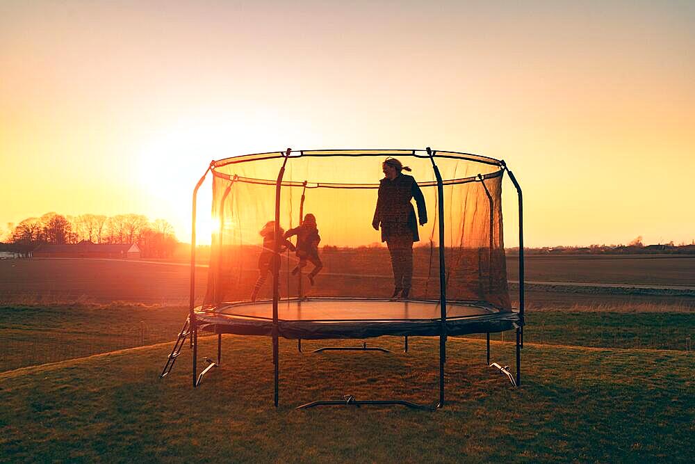 Trampoline on a lawn in the sunset with two kids and a young woman juping and playing
