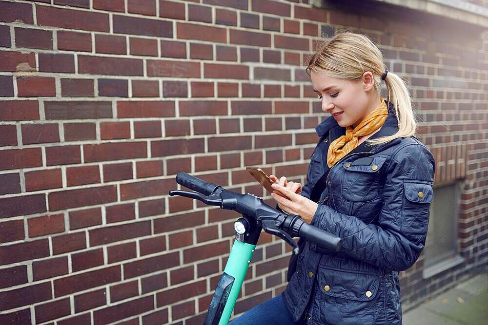 Trendy young blond woman in leather jacket standing at her e-scooter booking a ride, ecological transportation concept with copy space