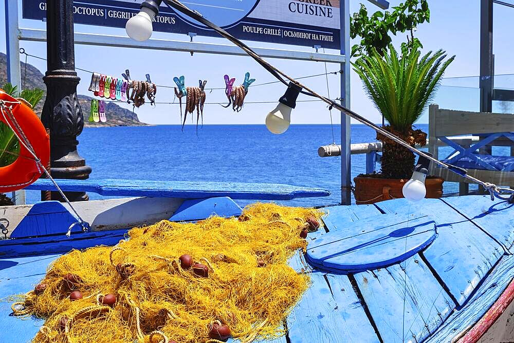 Outdoor installation of traditional fisherman's boat, fishing net and octopuses clipped to rope to dry in sun in selective focus and calm blue sea and typical Greek hills in background, Crete, Greece, Europe