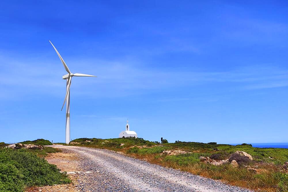 Single windmill turbine and small traditional Greek Orthodox chapel or church and road to them in colorful landscape on hilltop against blue sky with some cloulds on clear sunny summer day. Crete, Greece, Europe