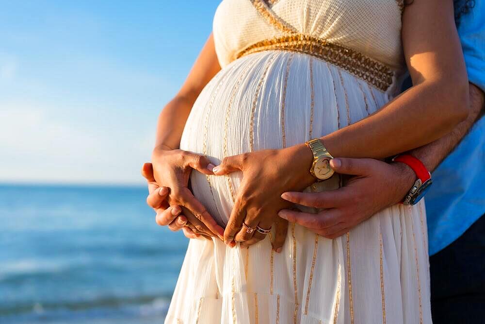 Pregnant maternity latin woman on the beach at sunset, maternity concept. Making the heart with the couple