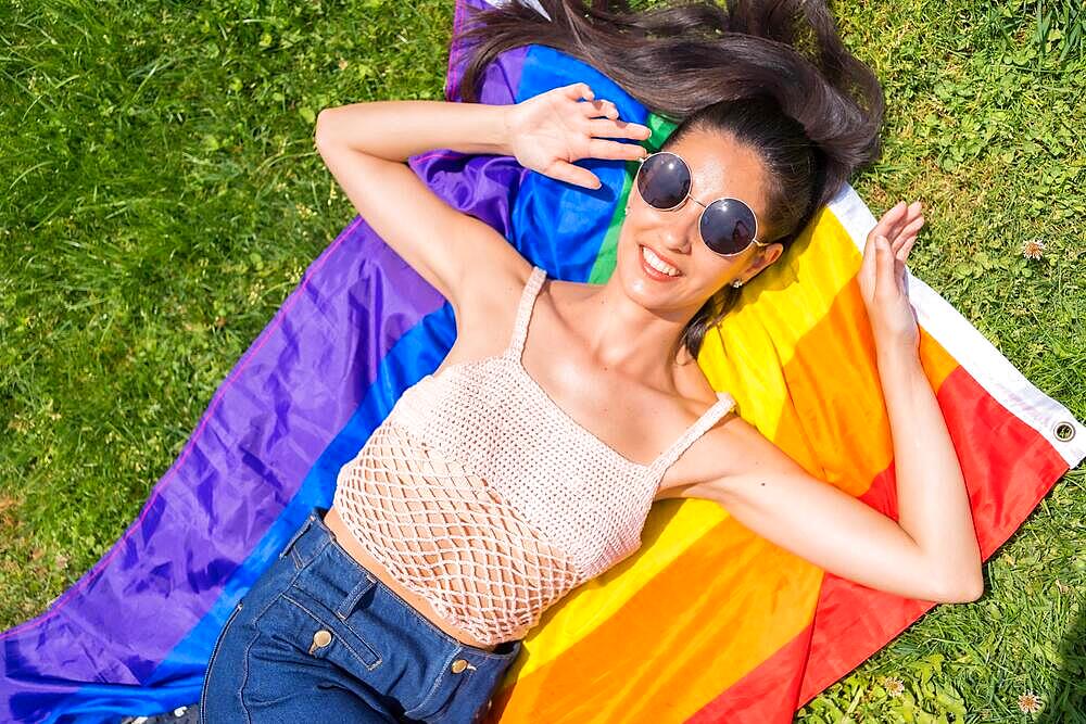 Caucasian brunette woman with a rainbow lgbt flag on the grass, lying down smiling