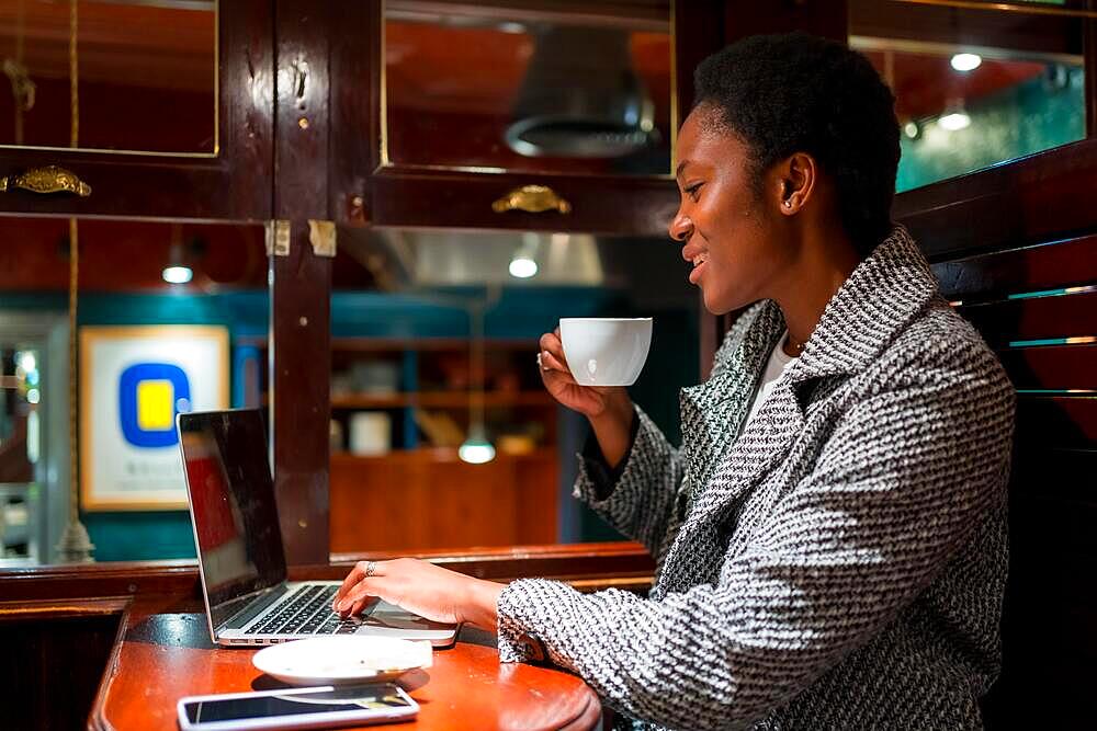 Business woman of African ethnicity in a coffee shop, working with a computer having a coffee