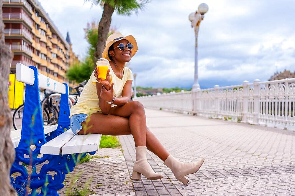 Portrait of black ethnic woman having a juice sitting on a bench in the city, tourist enjoying the summer