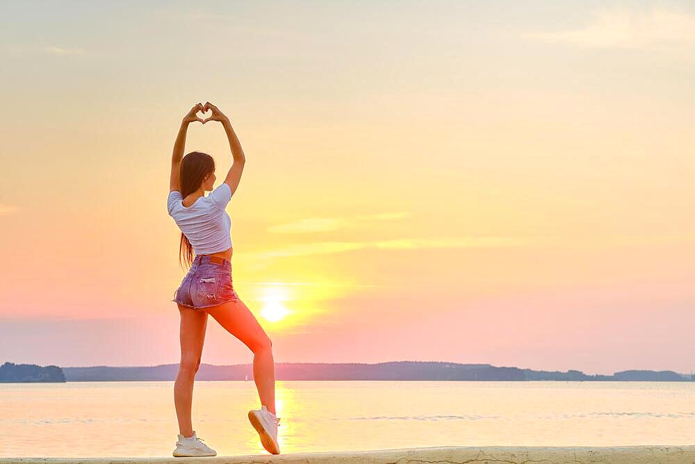 Woman raised her hands up and folded her palms in shape of heart against backdrop of sunset