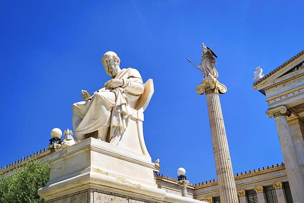 Marble statue of Plato, great ancient Greek philosopher, seated in chair and column or pillar with statue of Athena, ancient Greek goddess, patron of city, in background. Academy of Athens, Athens, Greece. Summer daylight sunshine. Blue sky