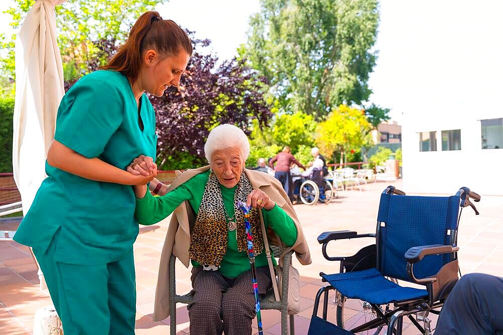 An elderly woman with the nurse in the wheelchair in the garden of a nursing home or retirement home, helping her to sit down