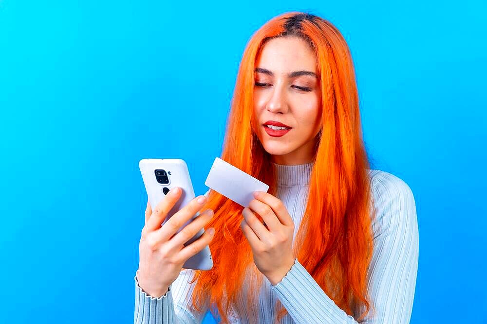 Redhead woman in studio photography online payment with mobile on a blue background