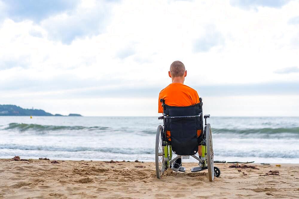 A disabled person on his back in a wheelchair on the beach in summer looking at the sea