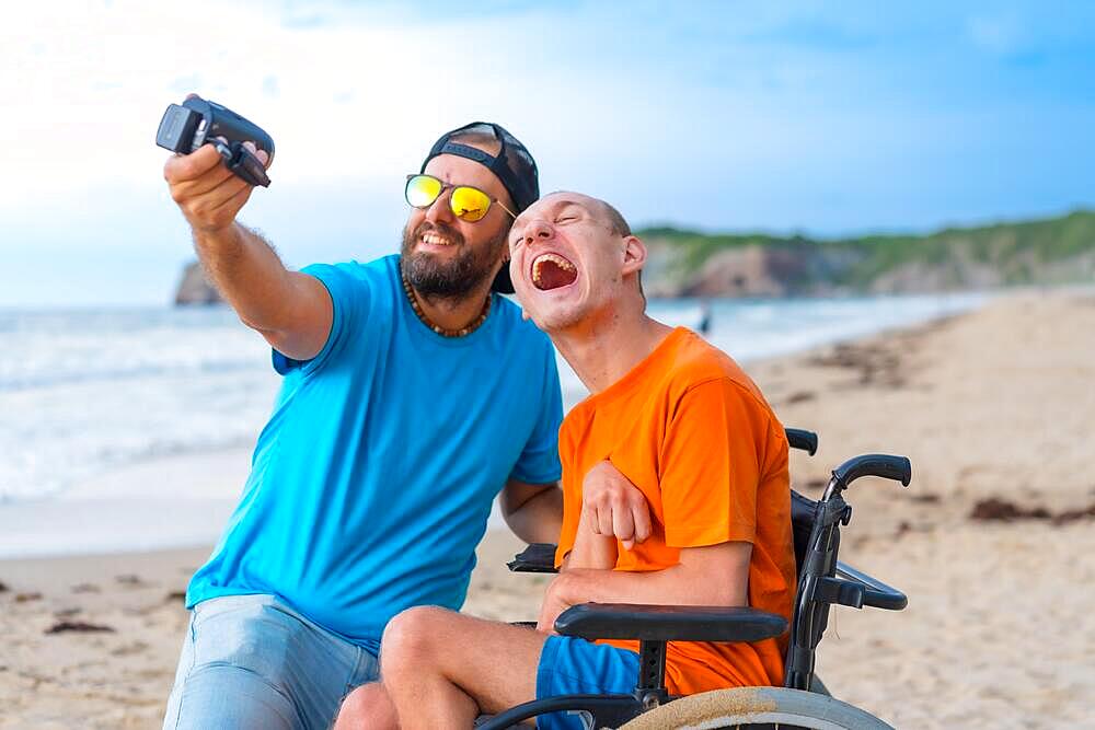 A disabled person in a wheelchair on the beach with a friend having fun taking a selfie