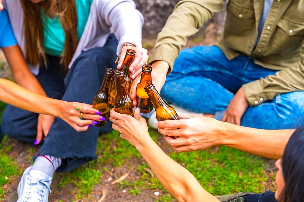 Group of multi-ethnic friends sitting in the city park talking next to a tree with bottles of beers