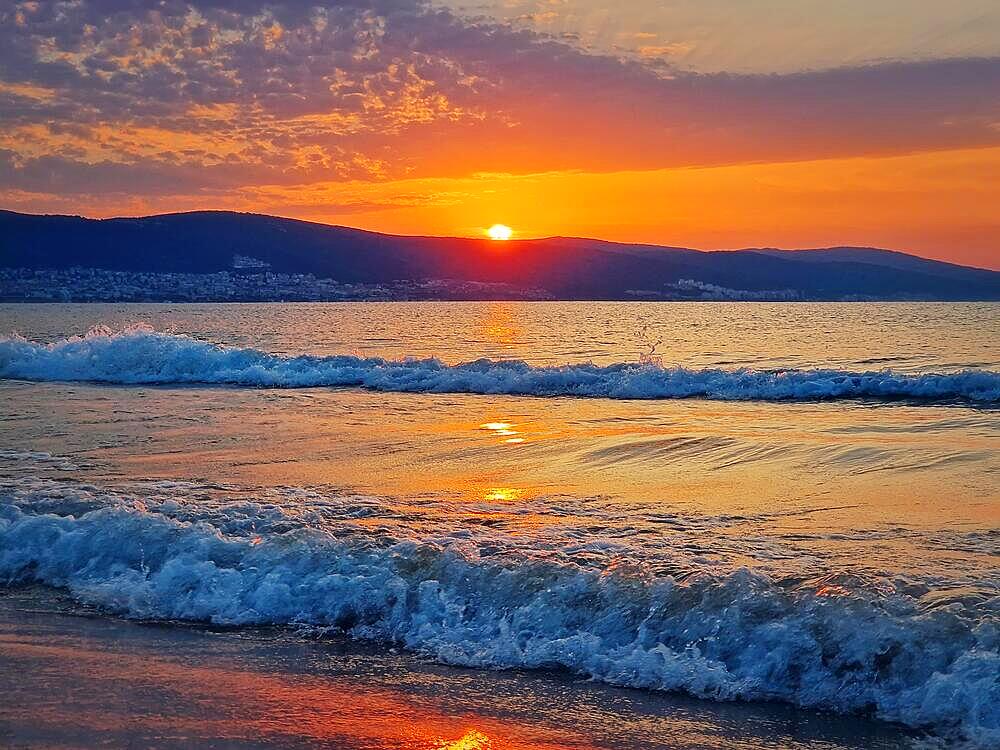 Beautiful sunrise at the Bulgarian coastline of Black Sea. Sunny beach resort, dawn scene with the sun rising up the hills on the horizon and foamy waves washing the sand shore. Summer vacation view