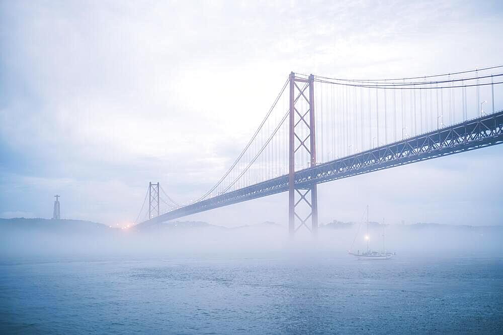 View of 25 de Abril Bridge famous tourist landmark of Lisbon connecting Lisboa and Almada in heavy fog mist wtih yacht boats passing under. Lisbon, Portugal, Europe