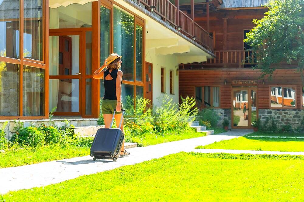 Tourist woman with a suitcase and hat on rural vacation arriving at a mountain cabin