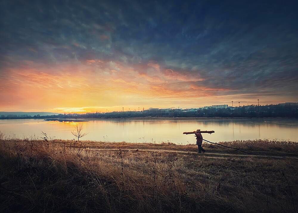 Person walking a country road carrying dry firewood. Peaceful sunset scene and wanderer silhouette on trail. Idyllic rural landscape