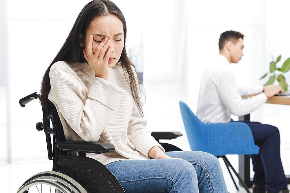 Worried young woman sitting wheel chair sitting front male colleague using laptop