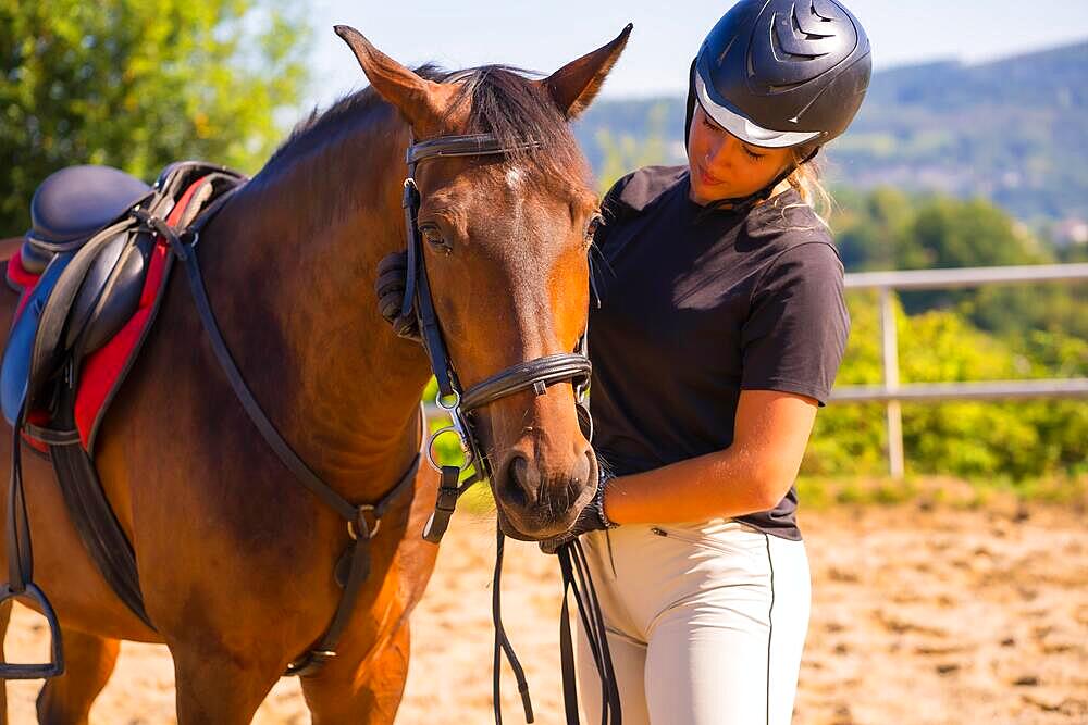 Posing of a Caucasian blonde girl on a horse caressing and pampering a brown horse, dressed in black rider with safety cap