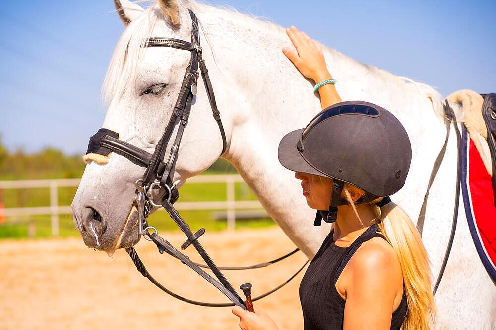 Caucasian blonde girl stroking a white horse on a horseback riding, dressed in black rider with safety hat