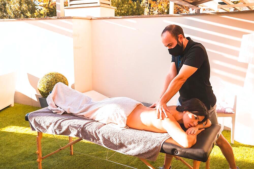 Massage at the client's home on the terrace with the sun in the background. Masseuse with face mask in the coronavirus pandemic