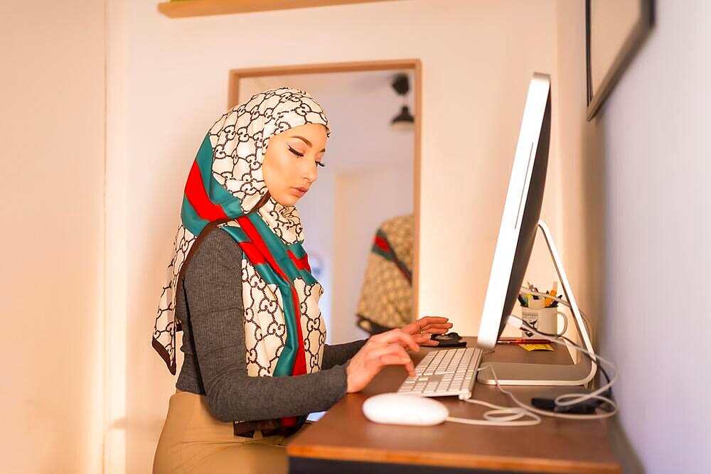 Veiled Arab girl at home computer, working from home. Modern Arab young woman with new technologies