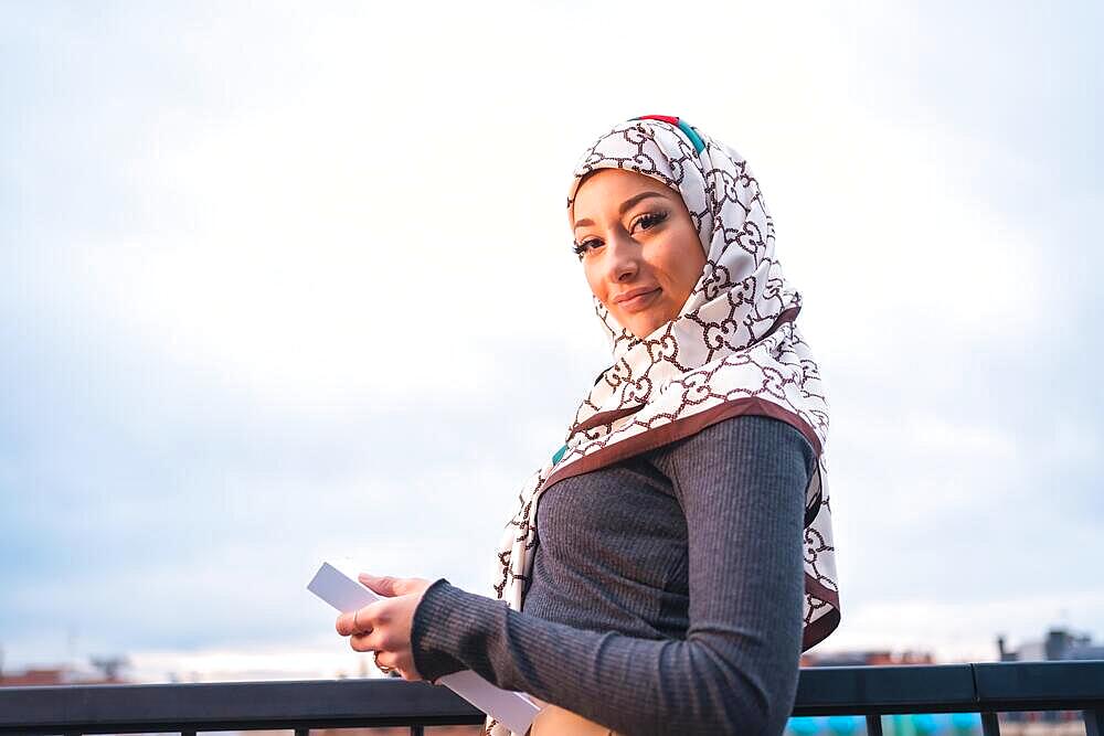 Portrait of an Arab girl with a white veil with a book on the terrace of a coffe shop, looking at the city from a balcony