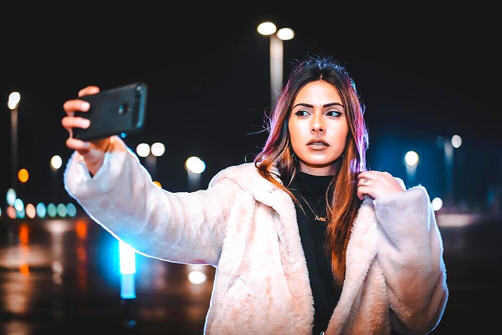 Caucasian brunette taking a selfie on the phone, wearing a pink wool jacket in an empty car park. Night urban session in the city