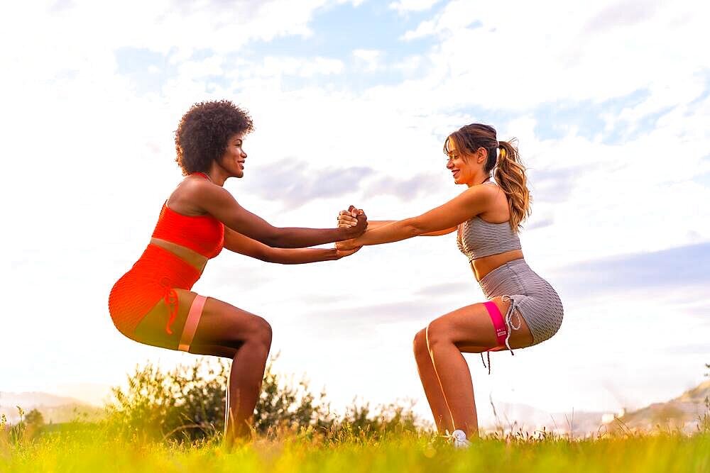 Caucasian blonde girl and dark-skinned girl with afro hair doing squat exercises in a park with the city in the background. Healthy life, fitness, fitness girls, gray and red sport outfits