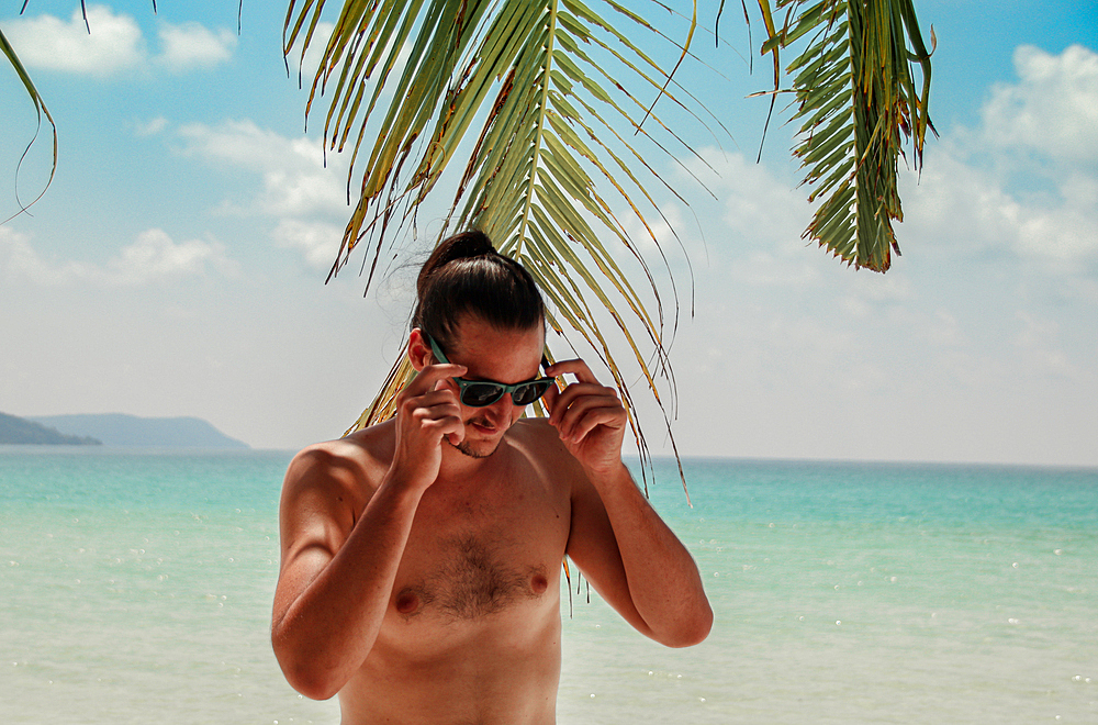 A shirtless man with a hair bun adjusts his sunglasses under palm leaves on a tropical beach with turquoise waters. Koh Rong Island, Cambodia, Asia