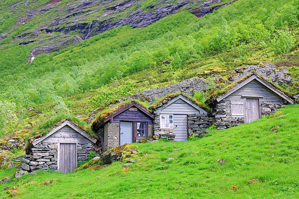 Three small rustic wooden cabins nestled in a green hilly landscape with grass and rocky mountains in the background, Alpine cabins, Nörangsdalen, Western Norway, Norway, Europe