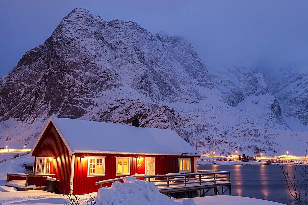 Red wooden house on fjord in front of snowy mountains, illuminated, twilight, winter, Reine, Moskenesoya, Lofoten, Norway, Europe