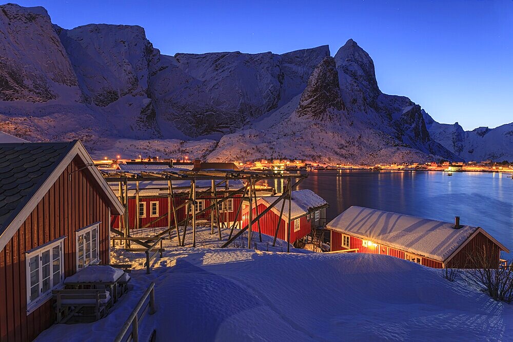 Red wooden houses on a fjord in front of snow-covered mountains, illuminated, twilight, winter, Reine, Moskenesoya, Lofoten, Norway, Europe