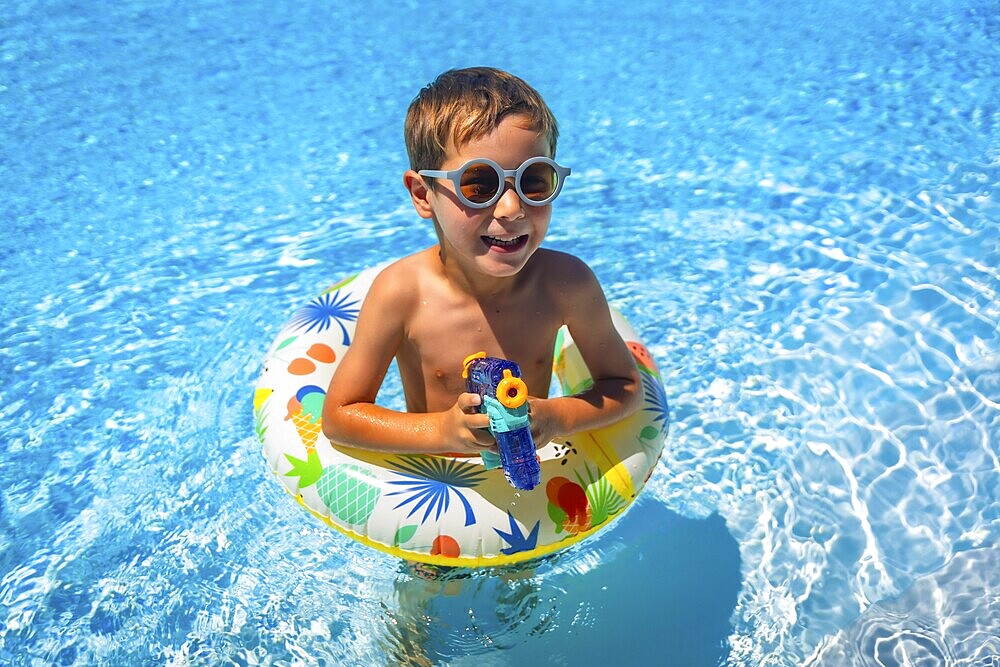 Boy aims with a water gun while playing with a float inside a blue swimming pool in summer