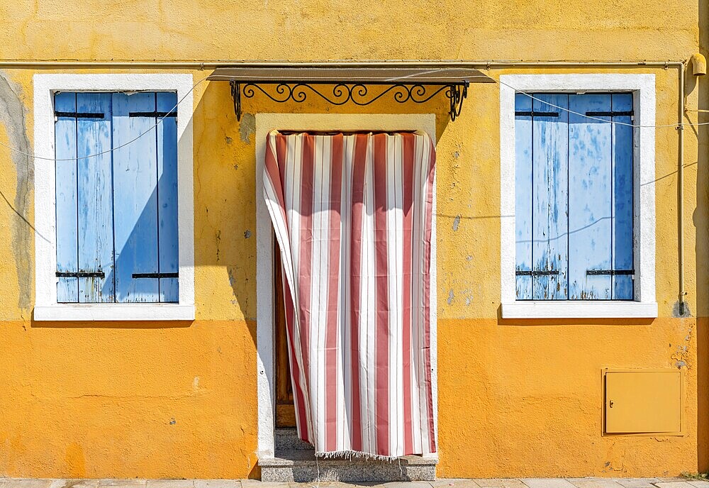 Yellow house facade with entrance door with red and white curtain and windows with blue shutters, colourful houses on the island of Burano, Venice, Veneto, Italy, Europe