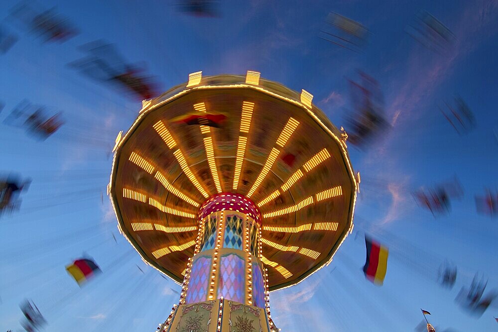 An illuminated carousel at dusk, spinning fast, in front of a blue sky with clouds and a Germany flag, Killiani Volksfest, Würzburg, Bavaria