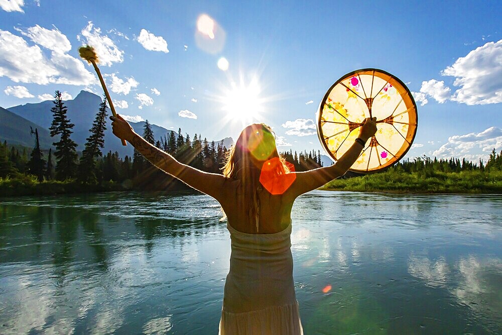 A close up and rear view of a shaman woman holding a sacred powwow drum towards sun and mountains during worship and healing ritual, with copy space