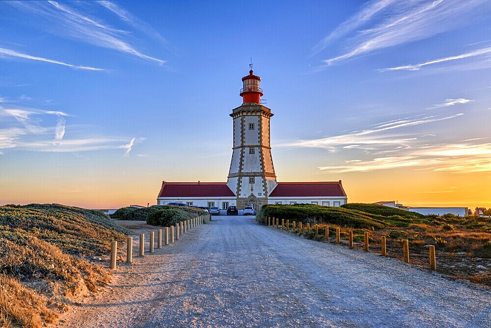 Beautiful of coastal lighthouse in warm colors of sunset, a clear path leading to it. Clear blue sky and some clouds, sun down over horizon, tranquil scene. Navigation beacon, safety for ships and small vessels, Atlantic ocean coast landscape