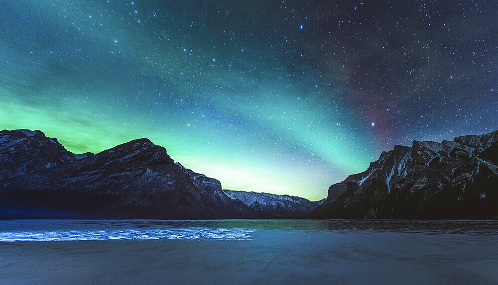 The image depicts the Northern Lights over mountains with a starry night sky and a serene lake in the foreground, Panorama shot, Canadian Rockies, Alberta, Canada, North America