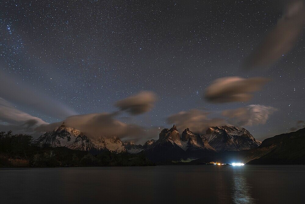 Night sky filled with stars over snow-capped mountains and a calm lake, with clouds and reflections creating a serene atmosphere, Torres Del Paine, Patagonia