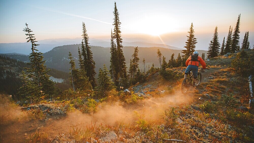 Mountain biker descending a forest trail at sunset, surrounded by trees and raising dust, Baldy Mountain, Nelson, BC, Canada, North America