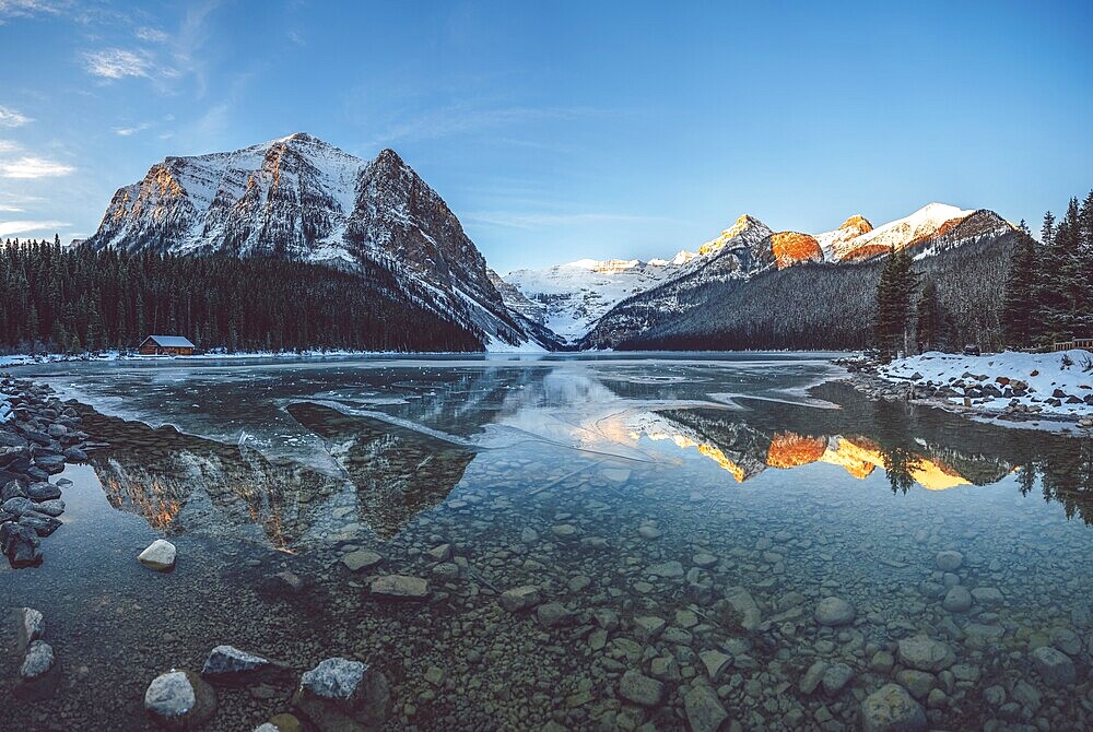 Peaceful lake reflecting snow-capped mountains and a cabin under a clear blue sky at sunrise, Lake Louise, Canadian Rockies