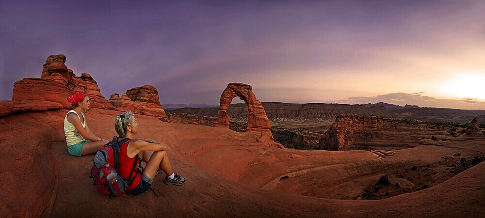 Hikers sitting at Delicate Arch natural stone arch at dusk, Arches-Nationalpark, near Moab, Utah, United States, North America