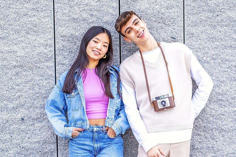 Frontal portrait of two multi-ethnic young friends leaning on a wall carrying a digital camera