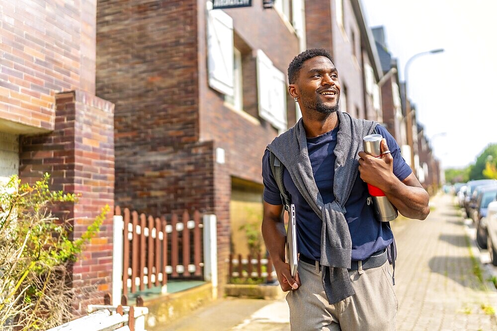 Confident young and handsome african man with casual clothes walking to go to work carrying a laptop and coffee reusable bottle
