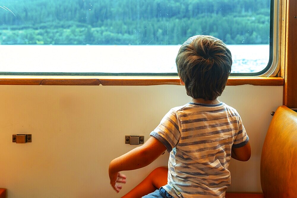 A young boy is sitting on a train window looking out at the water. The boy is wearing a striped shirt and shorts