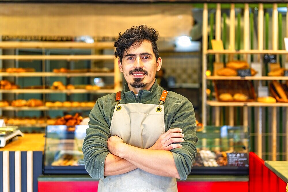 Formal portrait of a latin male young baker standing proud with arms crossed in a modern artisan bakery shop