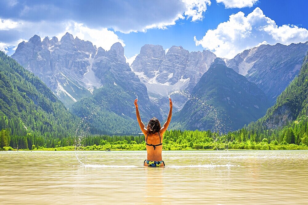 Young woman raising arms and splashing water in the famous lago di landro, also known as durrensee lake, in dolomites, italy, during summer vacation