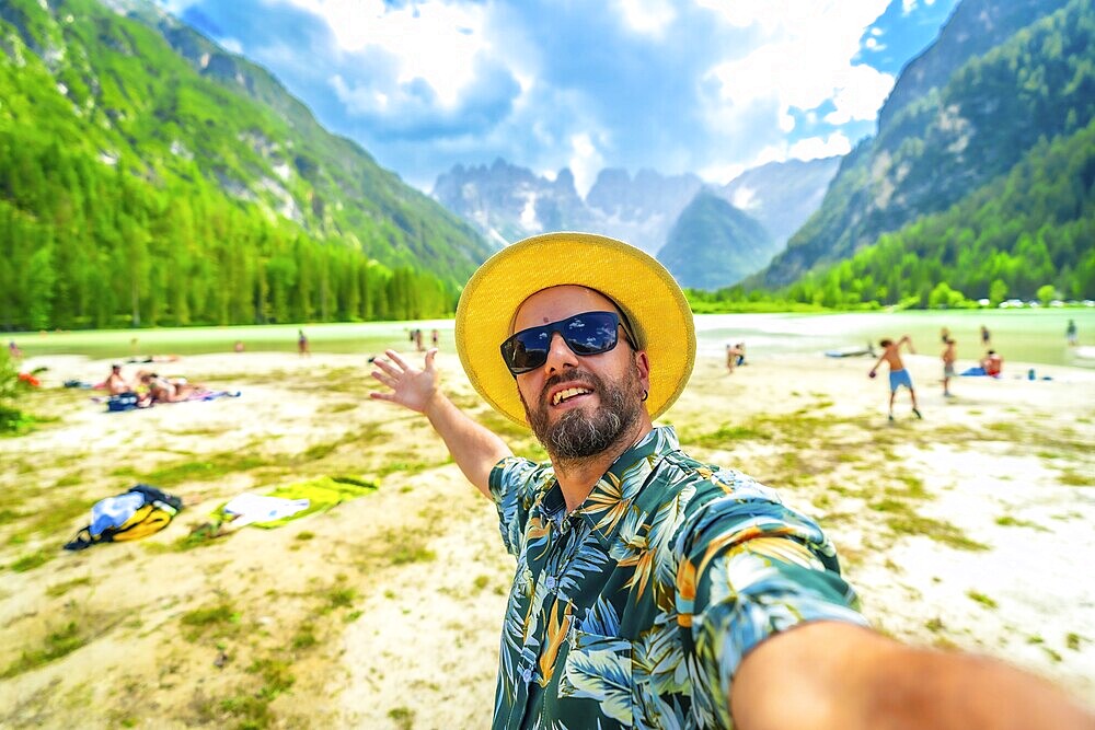 Happy man in a hawaiian shirt and straw hat taking a selfie at lago di landro, with tourists and tre cime di lavaredo in view