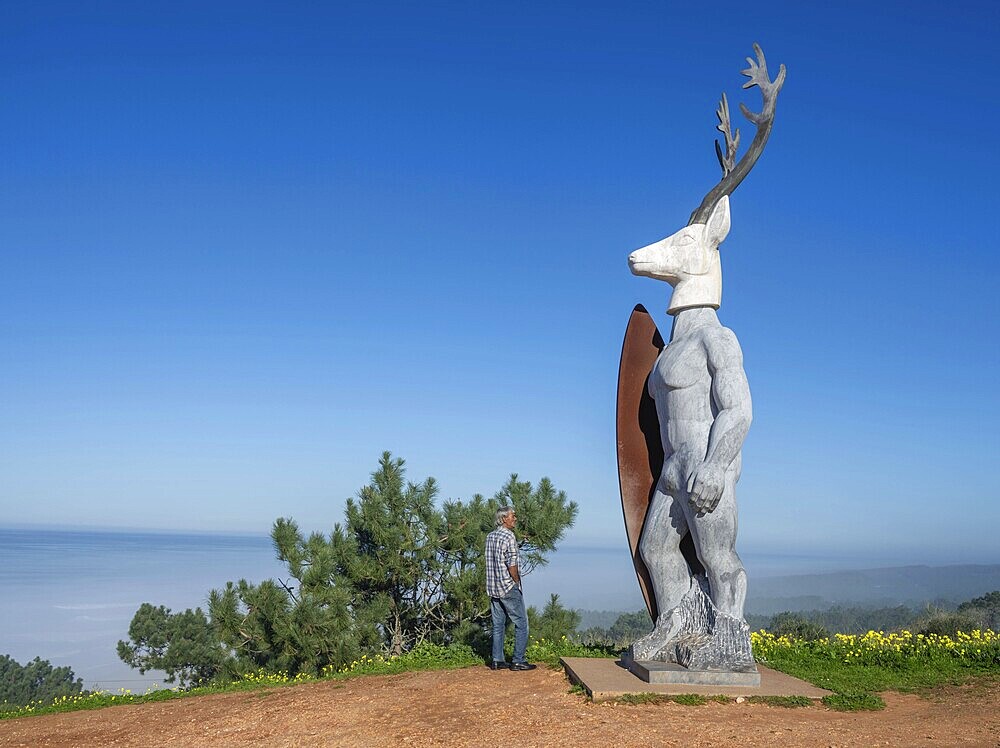 A man stands in front of a surfer statue with a deer head, Nazaré, Portugal, Europe